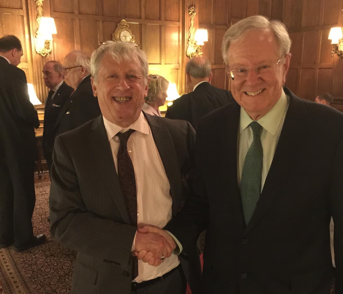 Ralph Benko, at left, shaking hands with Steve Forbes at right; both are smiling at the camera in a wood-panelled room with lit wall sconces and other guests behind them.