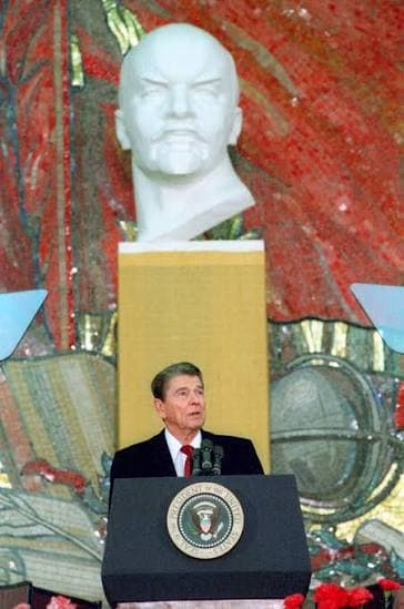President Reagan speaking from a lectern bearing the presidential seal at Moscow State University, beneath a monumental white bust of Lenin.